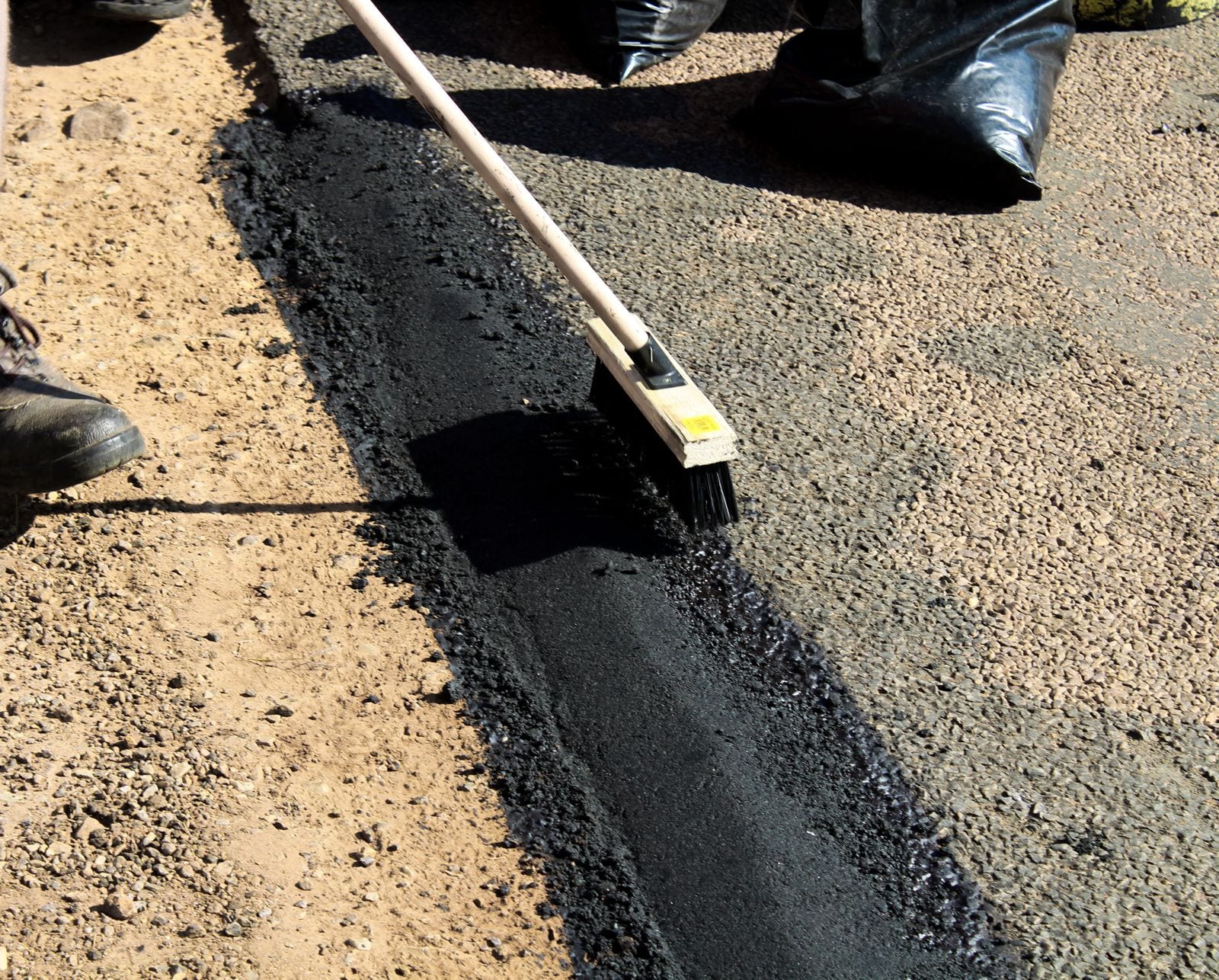 A brush and fresh black surfacing material beside an existing aggregate road surface