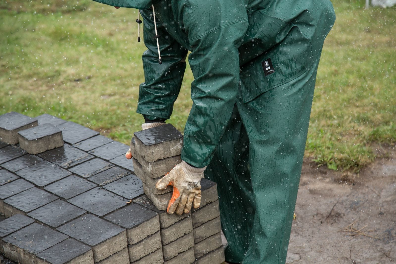 Gloved hands lifting grey paving blocks beside a partly paved surface